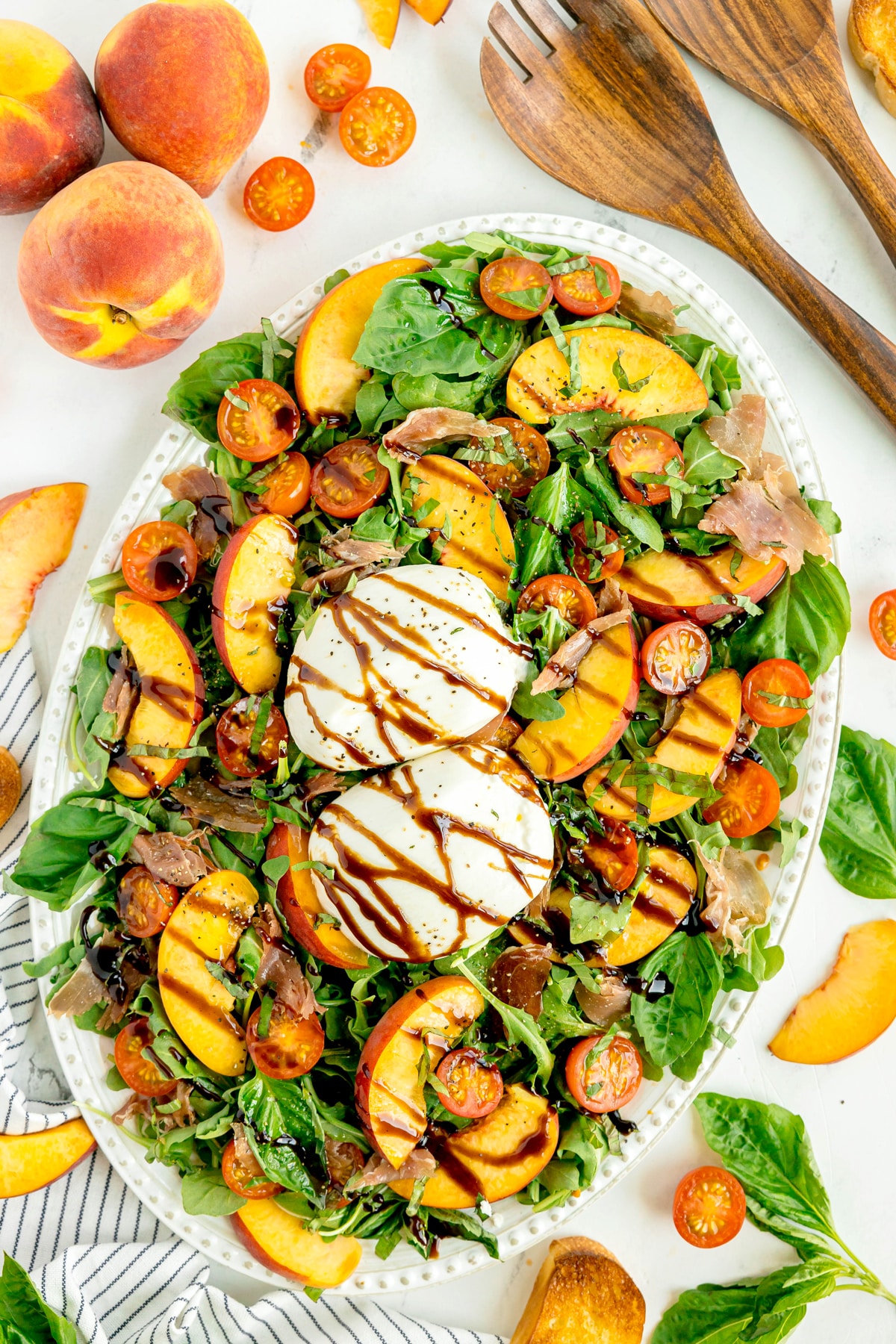 Oval platter of Burrata Peach Salad consist of mixed greens, fresh peaches, cherry tomatoes, two mozzarella balls, prosciutto, balsamic glaze, and fresh basil. Wooden utensils, peaches, and basil leaves surround the dish.