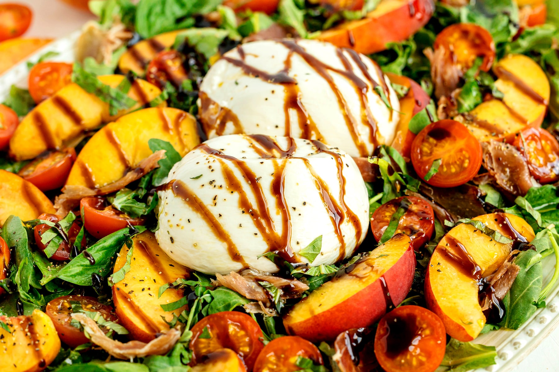 Burrata Peach Salad with arugula, sliced peaches, cherry tomatoes, prosciutto, and two burrata balls, drizzled with balsamic glaze.