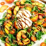 Arugula salad with sliced peaches, cherry tomatoes, burrata cheese, prosciutto, and balsamic glaze on a white platter.