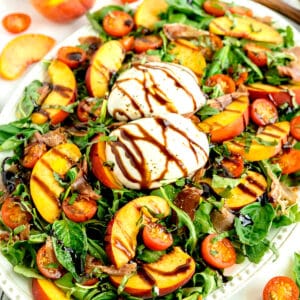 Arugula salad with sliced peaches, cherry tomatoes, burrata cheese, prosciutto, and balsamic glaze on a white platter.