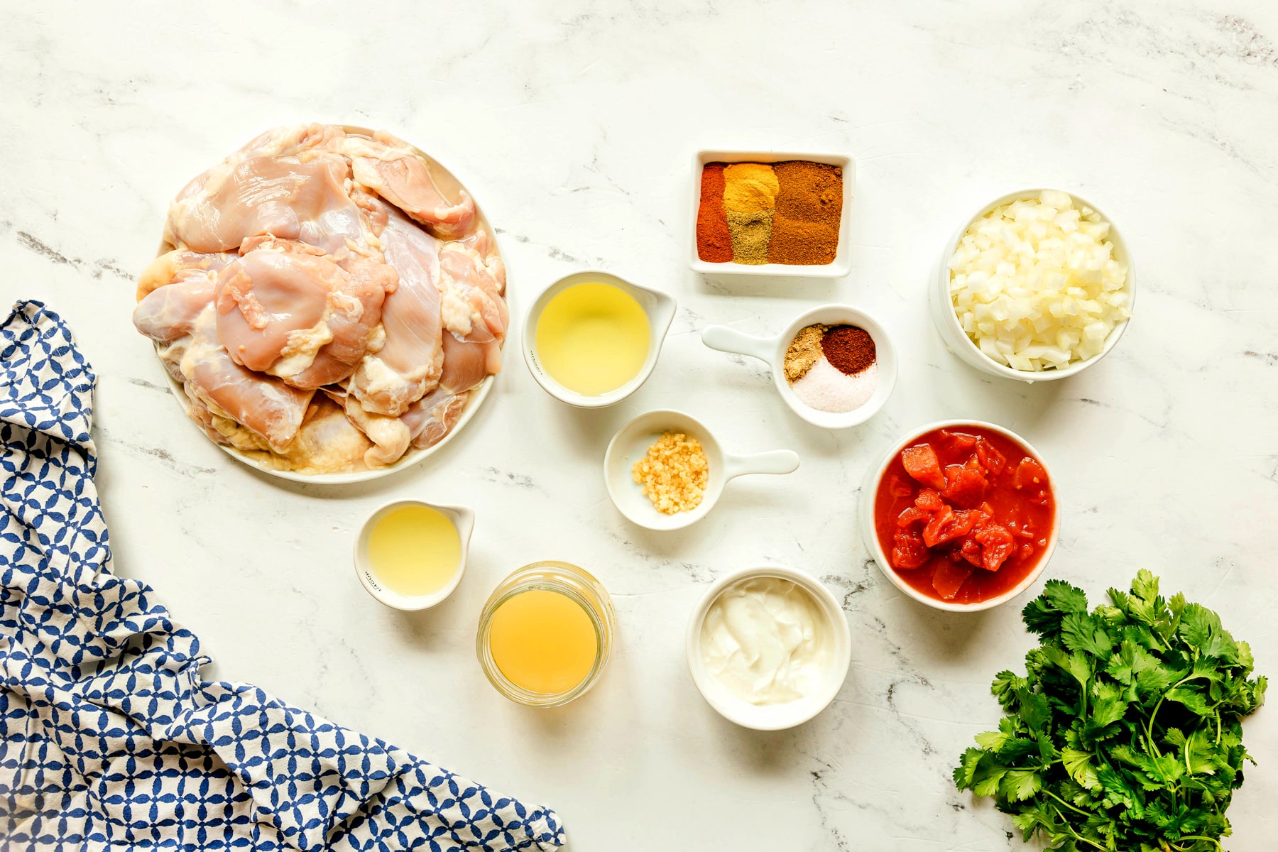Ingredients for Chicken Masala Recipe include chicken thighs, vegetable oil, garam masala, smoked paprika, cumin, turmeric, chili powder, onion, minced ginger, garlic, salt, diced tomatoes, chicken stock, lemon juice, yogurt, and cilantro, arranged on a marble countertop.