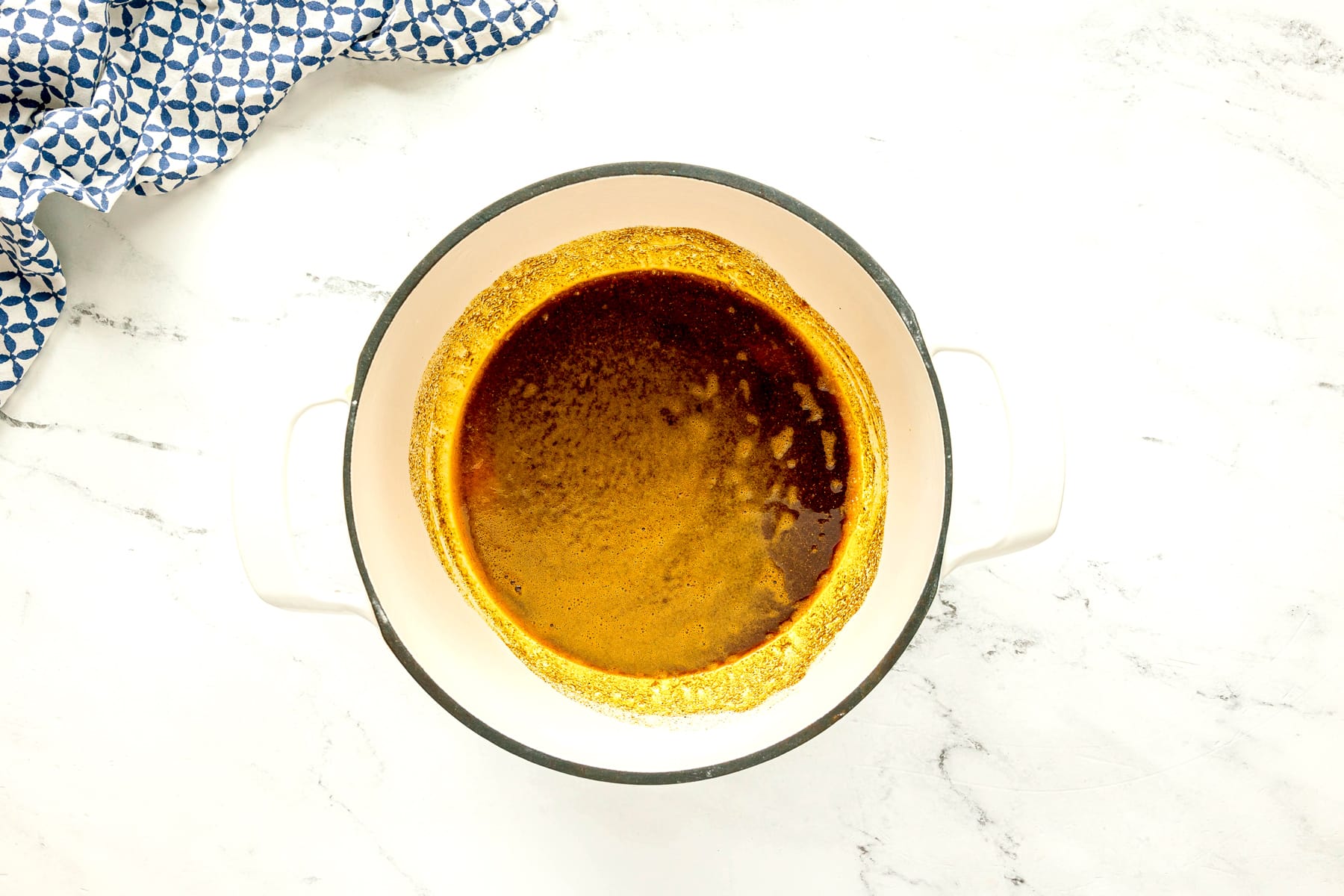 First step in preparing Chicken Masala Recipe is to use a white pot. It has garam masala, paprika, cumin, turmeric, and chili powder and toast and sits on a white marble surface next to a blue patterned cloth.