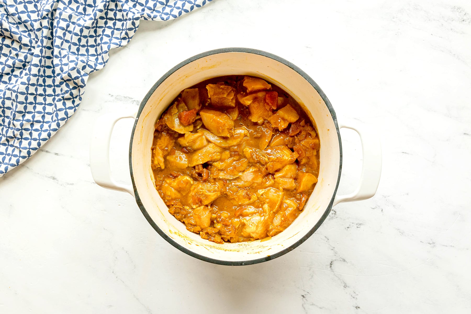 A white pot filled with cooked chicken in Chicken Masala sauce sits on a white marble surface next to a blue and white patterned cloth.