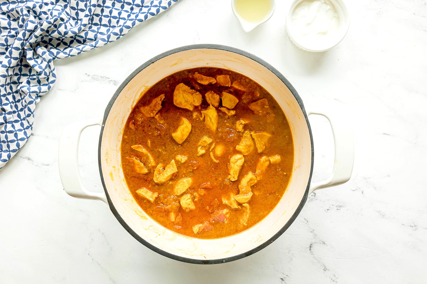 A white pot containing chicken pieces in Chicken Masala with added water, placed on a white surface next to a patterned cloth and two small bowls with liquid and white sauce.
