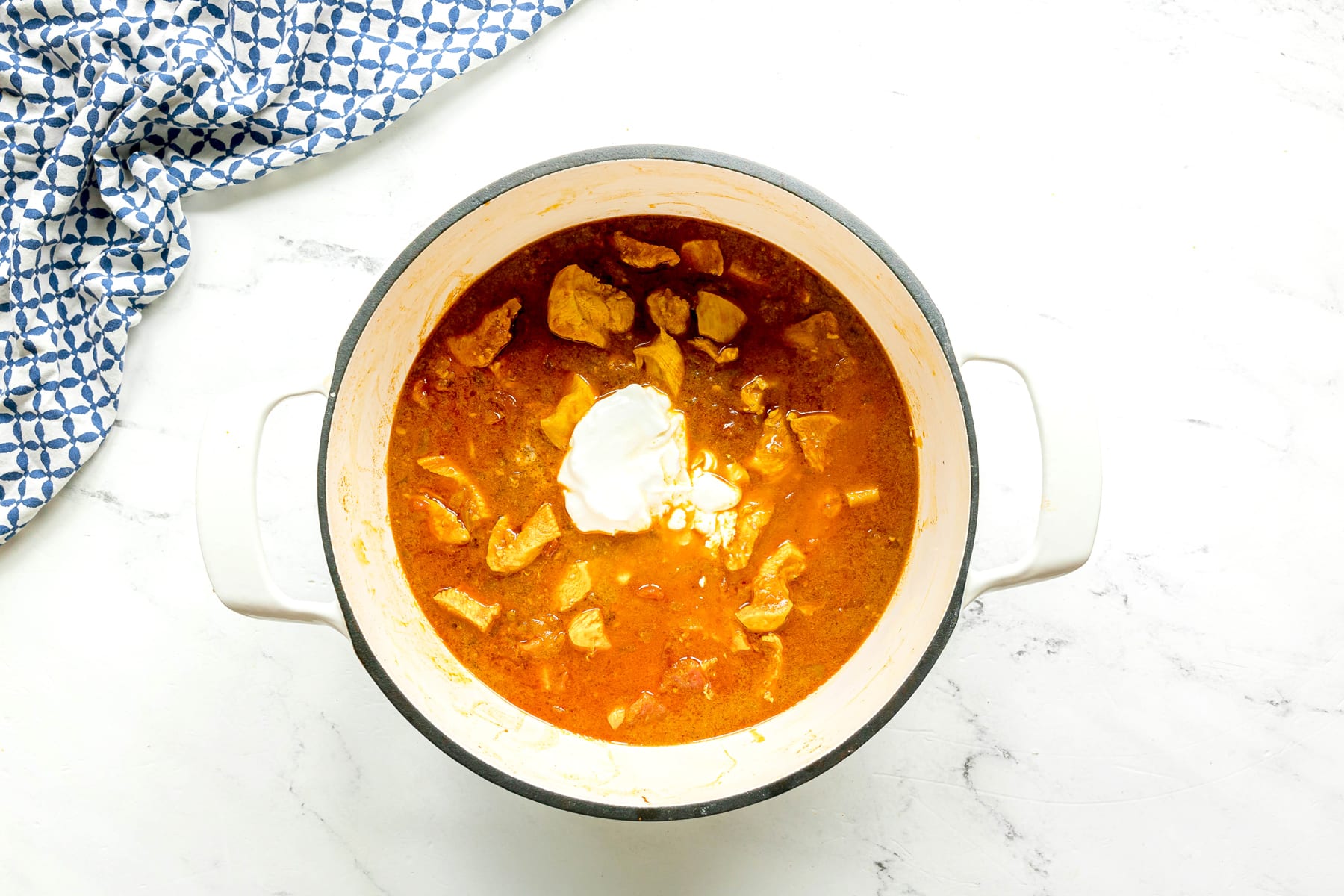 A white pot filled with a Chicken Masala mixture, containing meat chunks, topped with a dollop of yogurt, sits on a white surface next to a blue and white patterned cloth.
