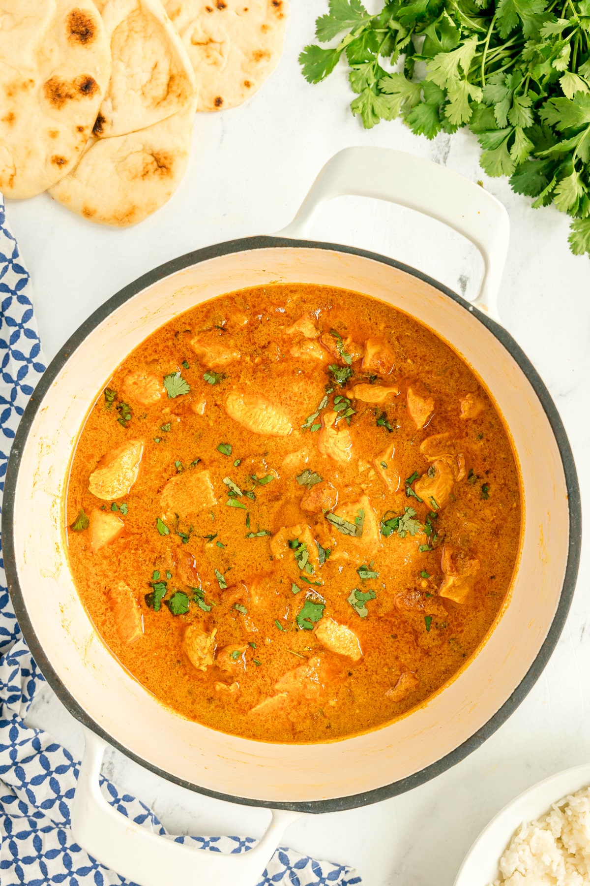 A pot of Chicken Masala Recipe garnished with cilantro, shown with naan bread, fresh cilantro, and a bowl of rice on a white surface.