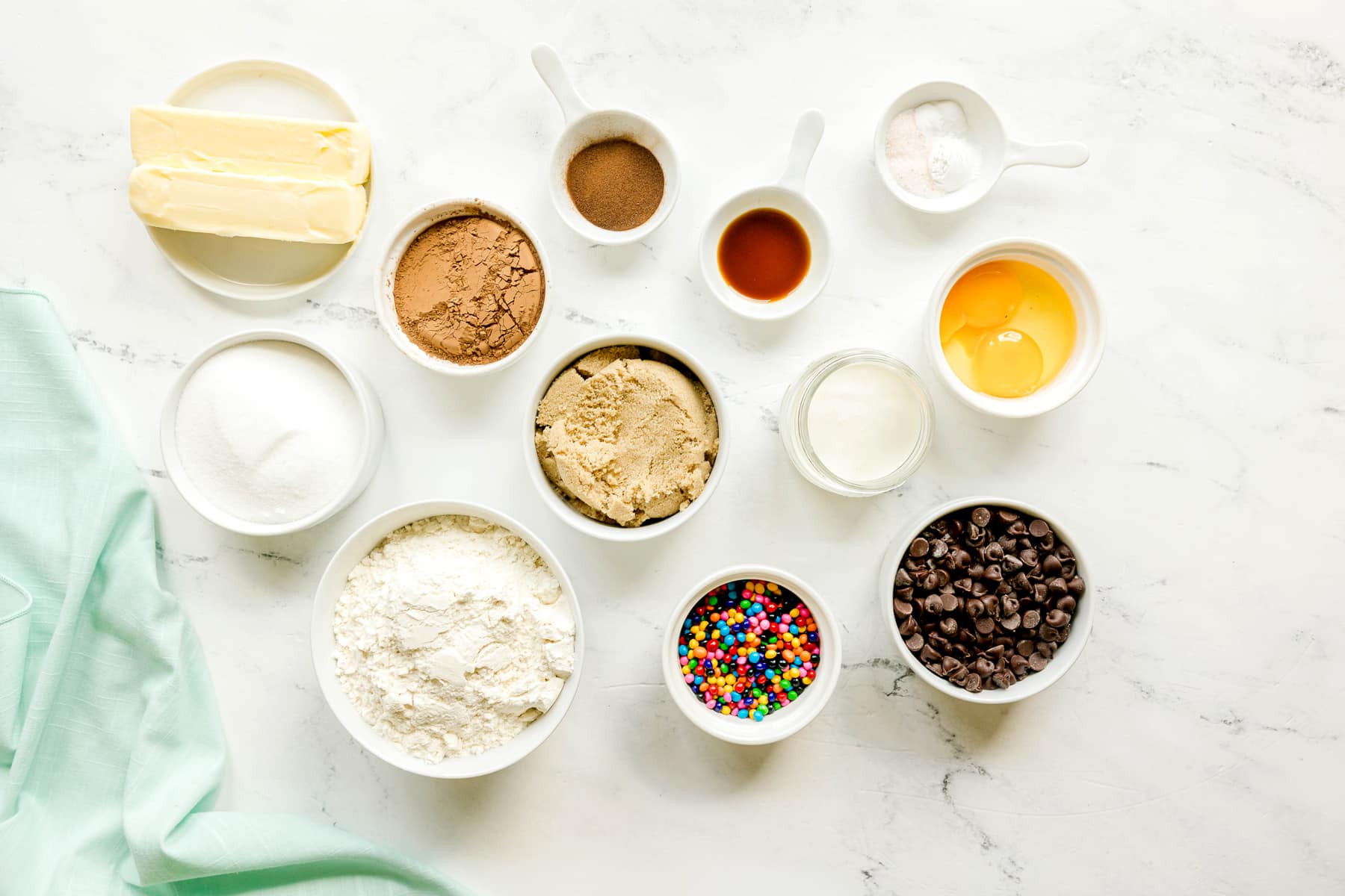 Various baking ingredients for Cosmic Cookies, including butter, cocoa powder, instant espresso powder, granulated sugar, brown sugar, egg and egg yolk, vanilla extract, all-purpose flour, baking powder, baking soda and salt, arranged on a marble surface.