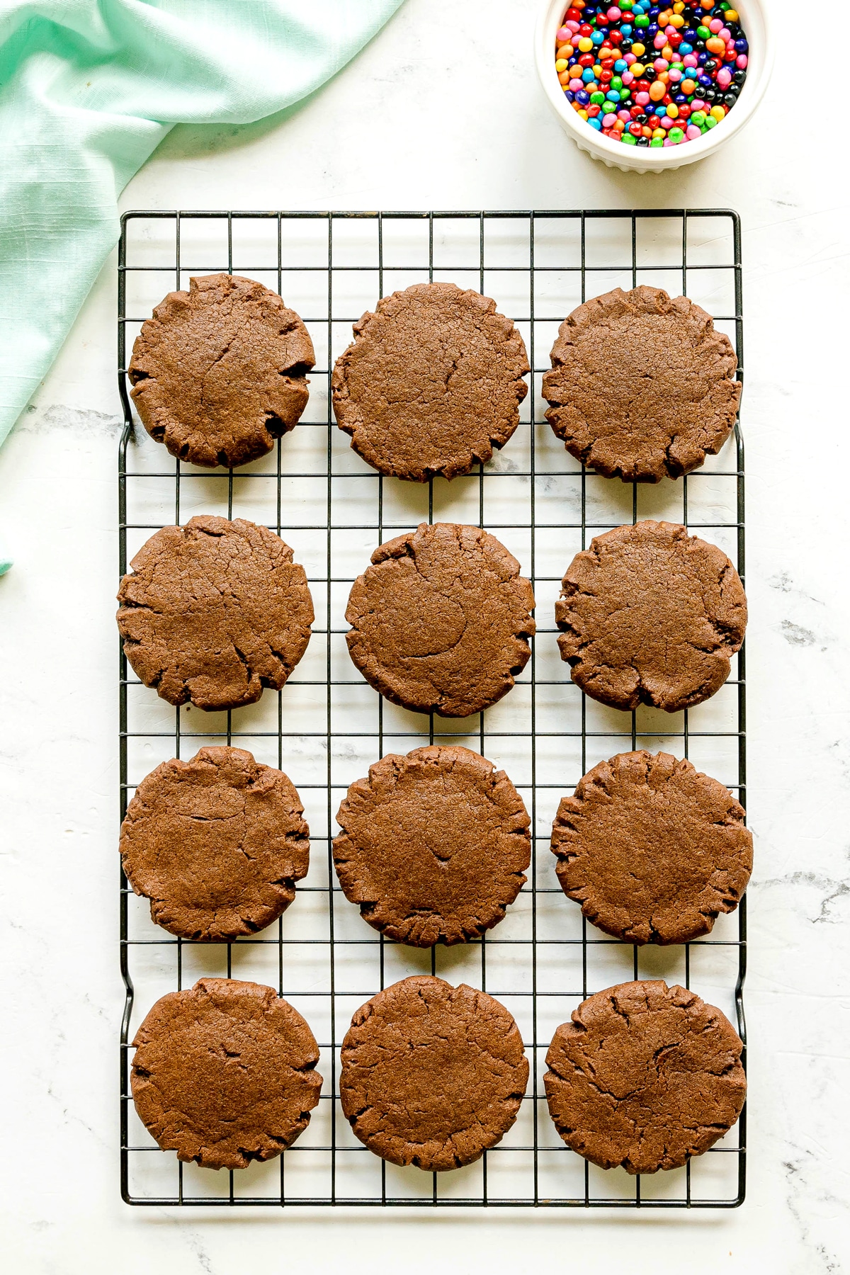 Twelve chocolate cookies are cooling on a wire rack, with a bowl of colorful candy-coated chocolates in the upper right corner.