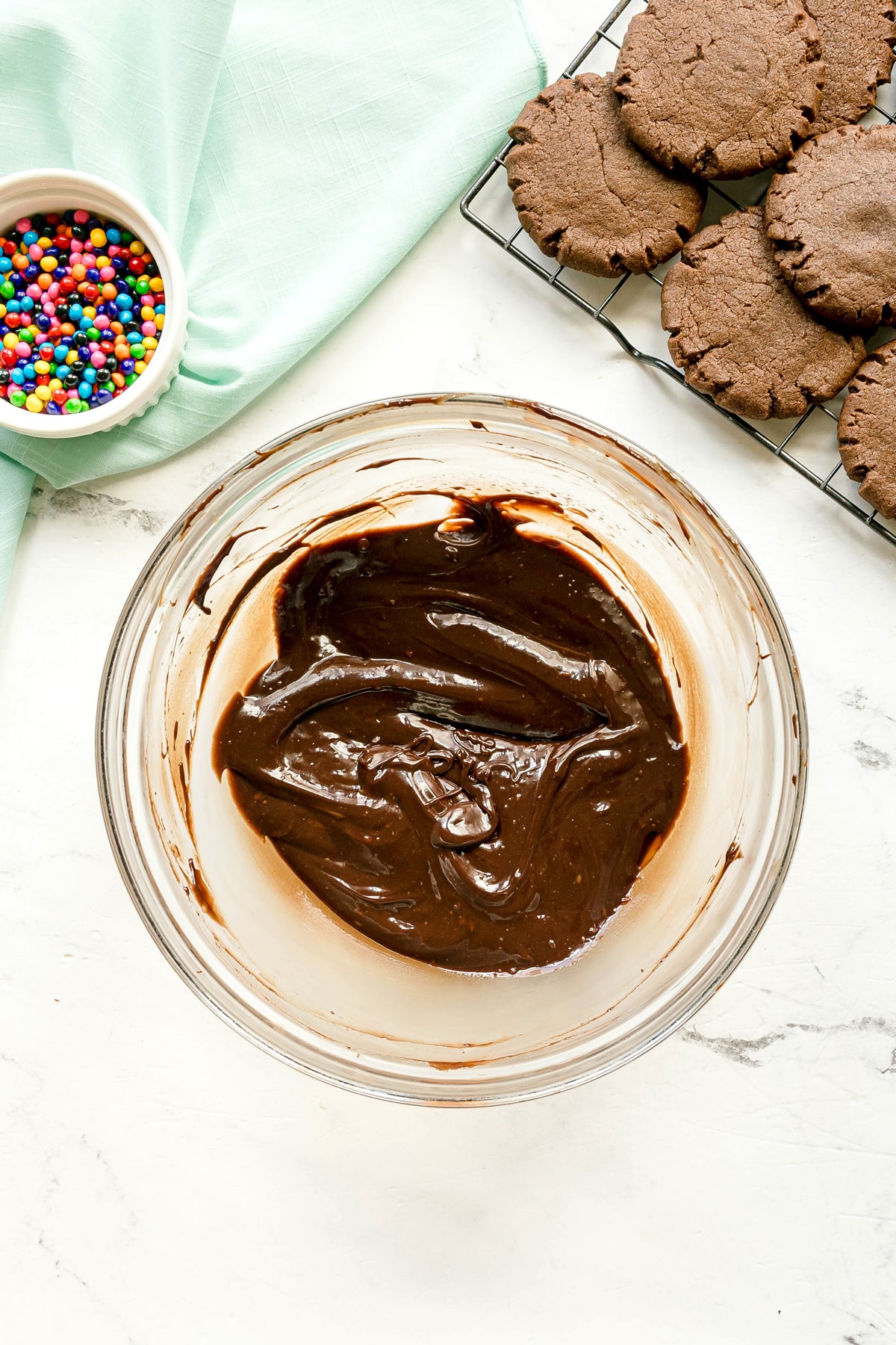 A glass bowl of chocolate batter sits on a white surface near a rack of chocolate cookies and a small bowl of colorful sprinkles.