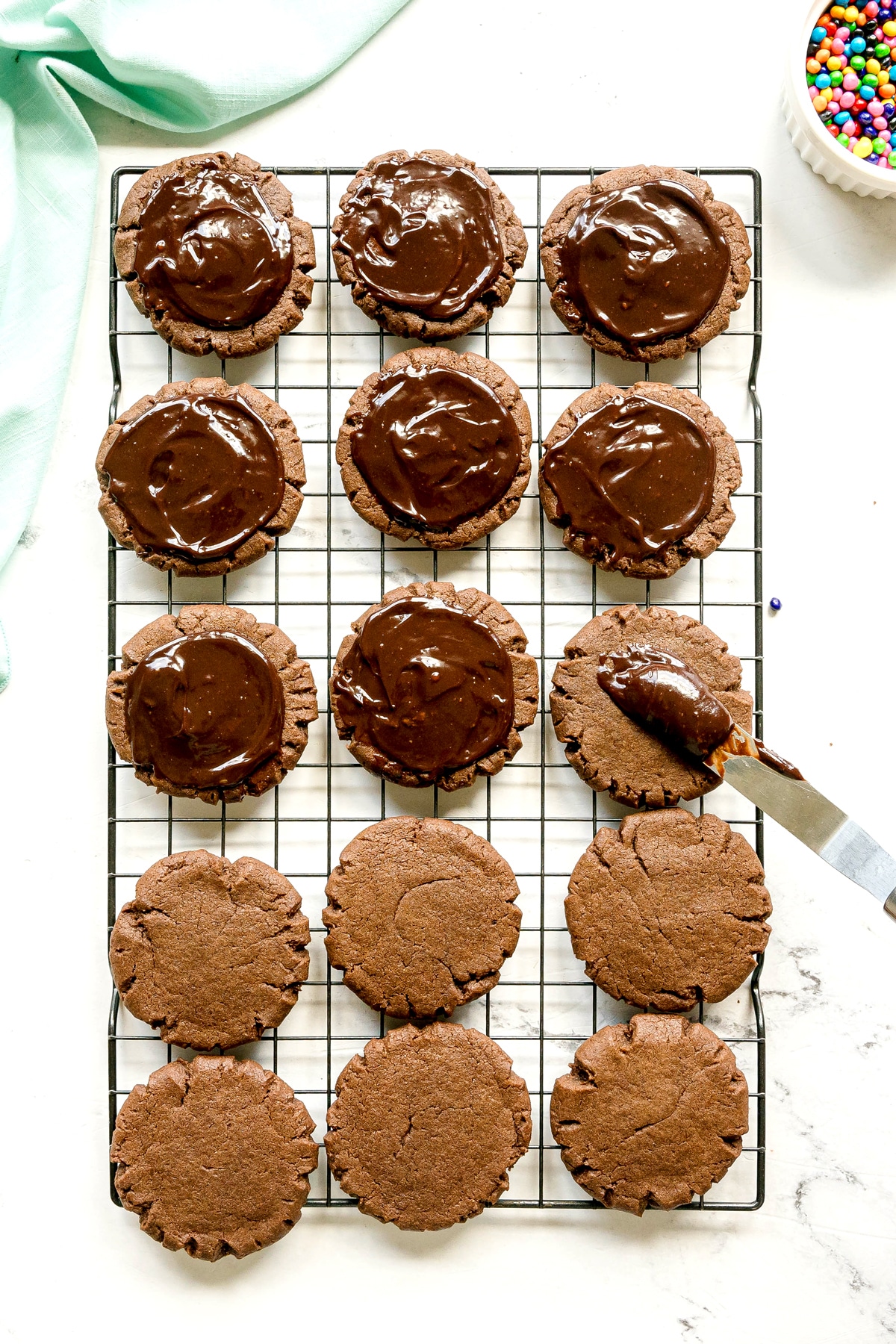A cooling rack with fifteen chocolate cookies, some topped with chocolate frosting. A spatula with frosting is beside one cookie being iced. A bowl of colorful candies is in the corner.