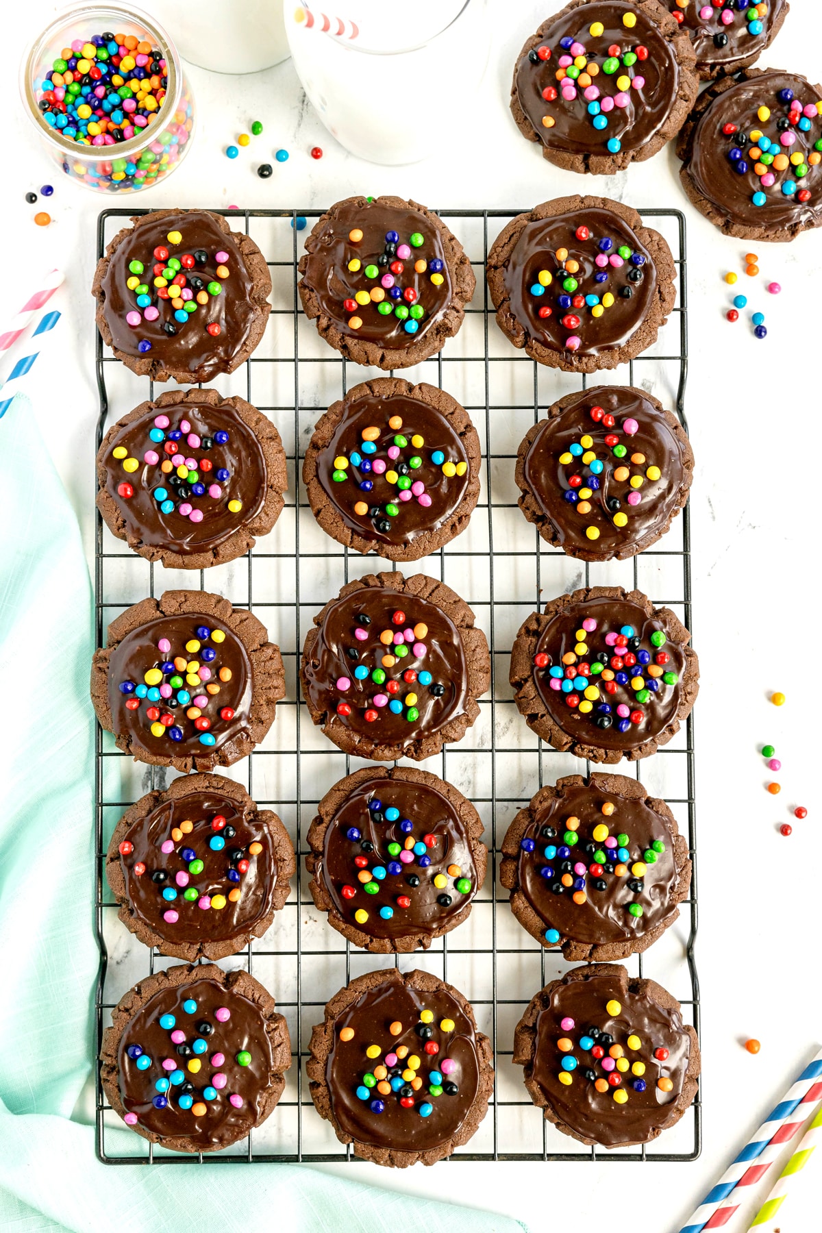 Sixteen Cosmic cookies with chocolate icing and colorful candy sprinkles arranged on a cooling rack, with extra cookies, milk, and straws visible around the edges.