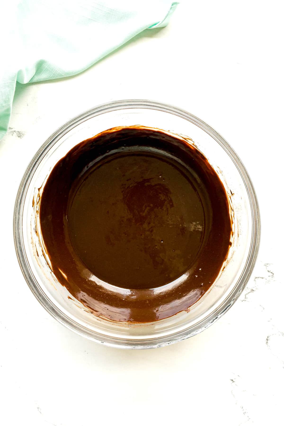 A glass bowl filled with smooth melted butter, cocoa powder and espresso powder sits on a white surface with a light green cloth nearby for Cosmic Cookies.