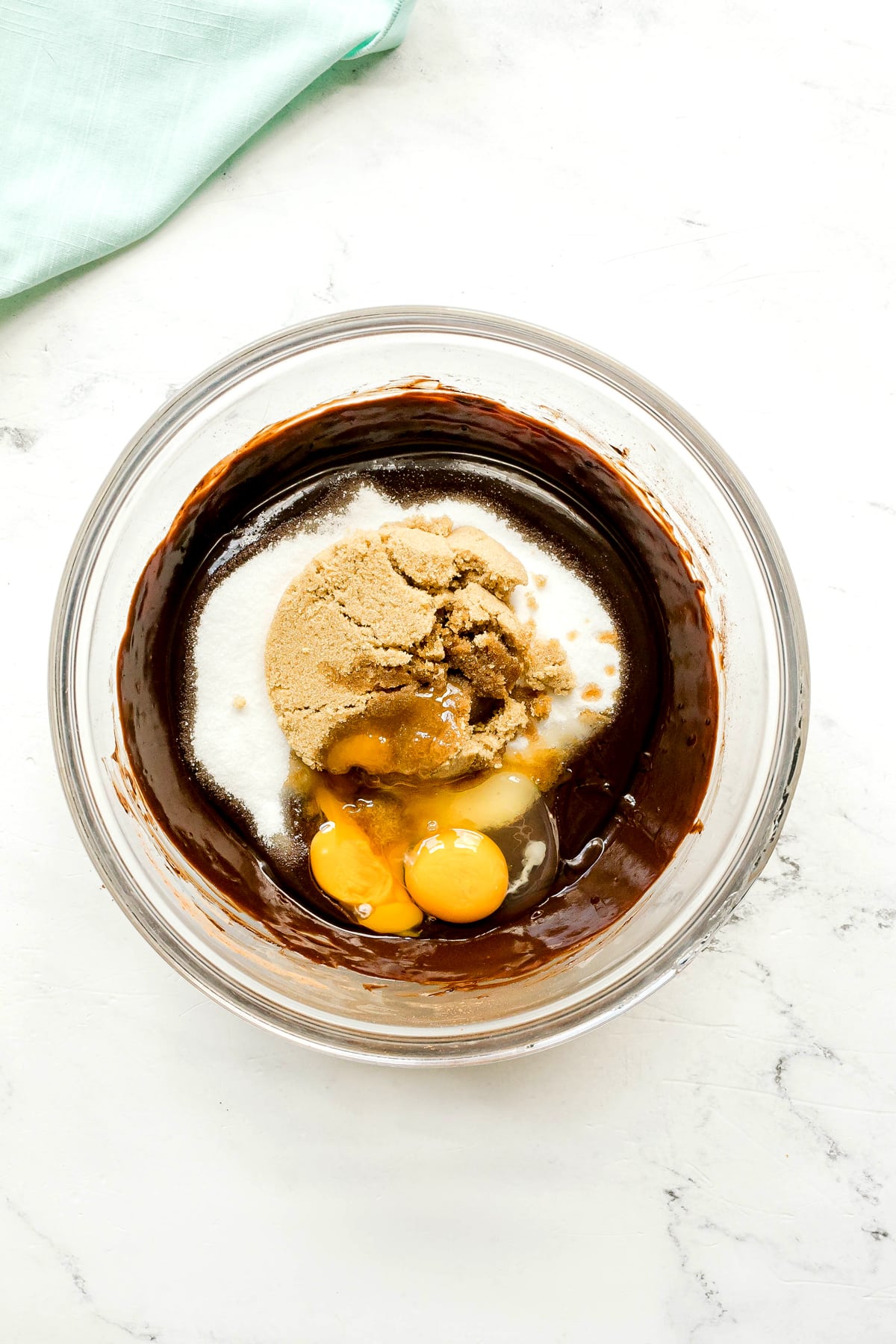 Next step in preparing Cosmic cookies is to use a clear glass bowl and add chocolate batter, brown sugar, egg, egg yolk and vanilla. A folded light green cloth is partially visible in the corner.