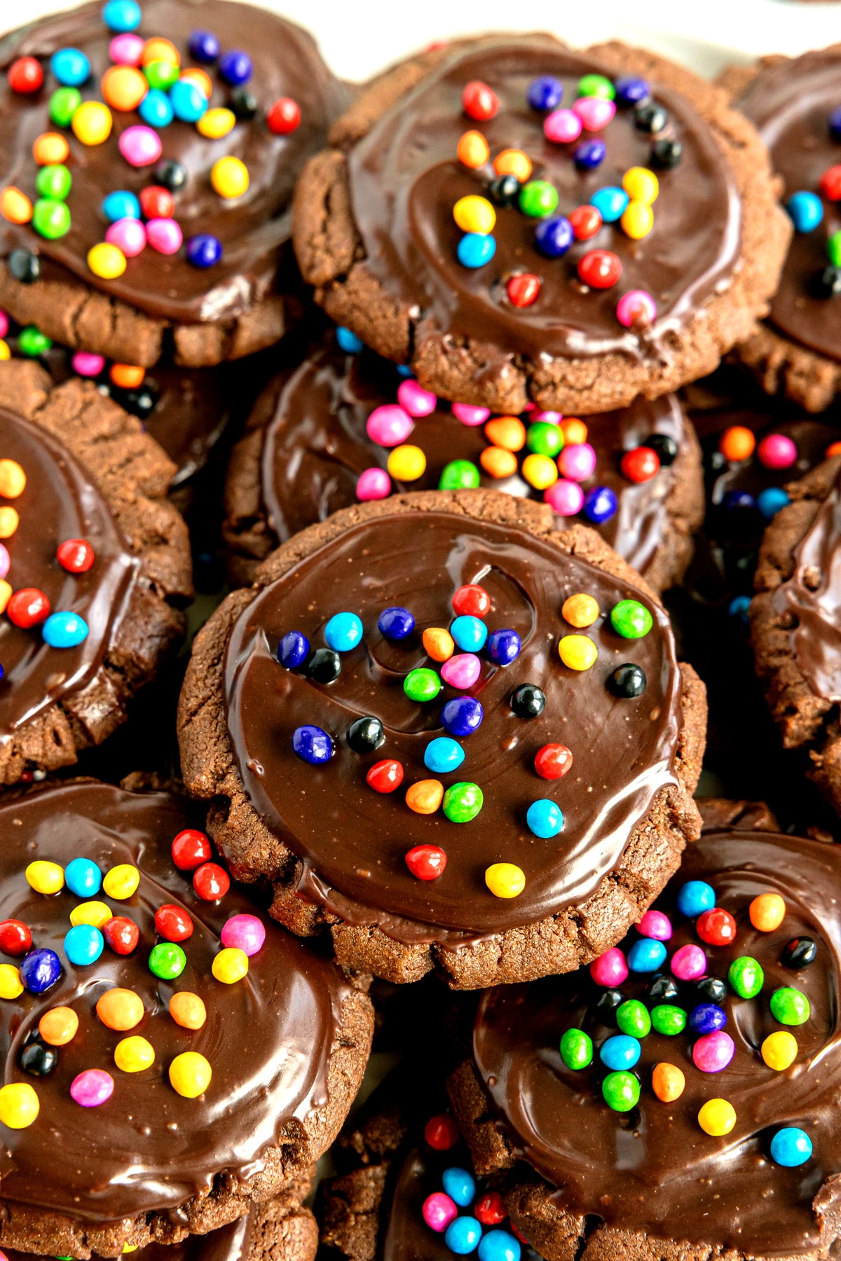 Cosmic cookies topped with a layer of glossy chocolate frosting and colorful round candy sprinkles, closely stacked together.
