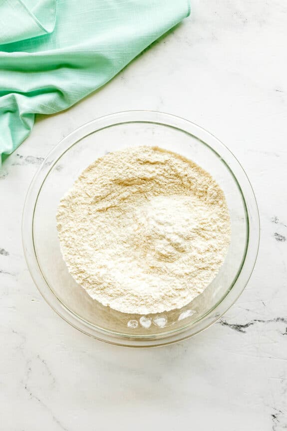 A clear glass bowl filled with flour, baking powder, baking soda, and salt for Cosmic cookies, sits on a marble surface, next to a folded light green cloth.