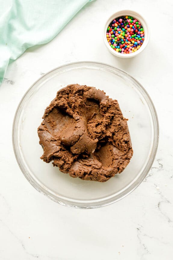 A glass bowl of Cosmic cookie dough sits on a white surface next to a small bowl of colorful round sprinkles.