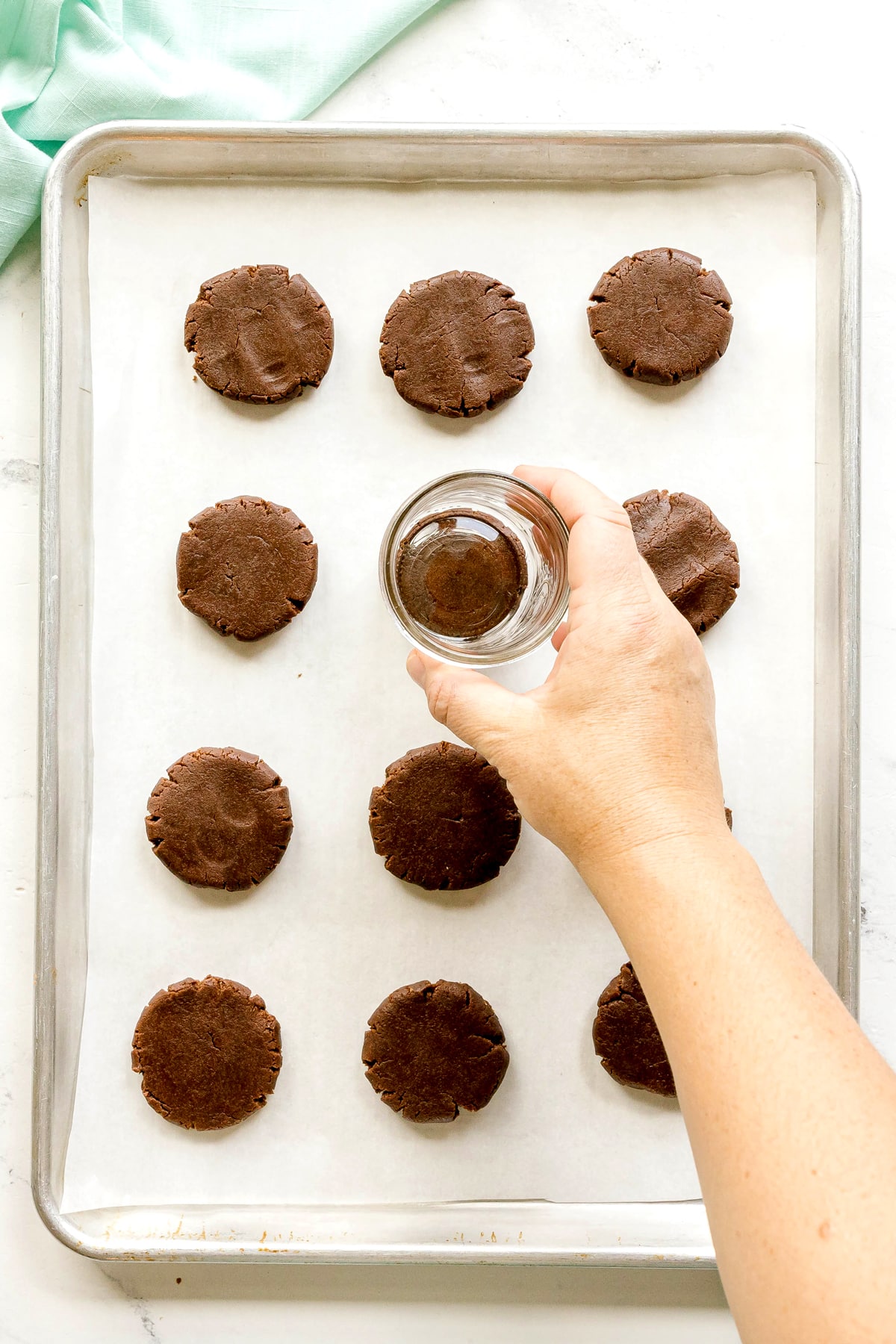 A hand presses a small glass onto Cosmic Cookie dough rounds arranged on a parchment-lined baking sheet.