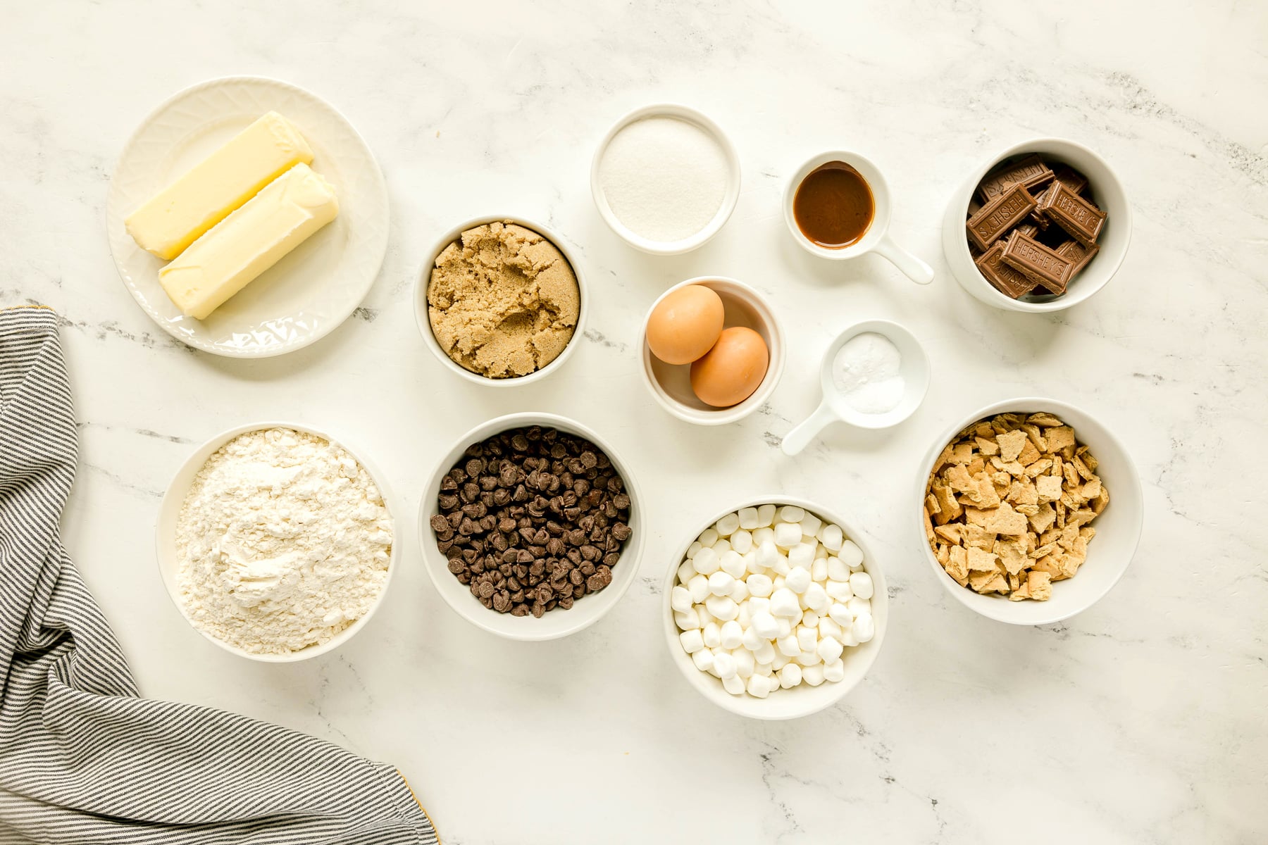 Various baking ingredients for S'mores Cookies include eggs, vanilla extract, all-purpose flour, baking soda, salt, semi-sweet chocolate chips, mini marshmallows, graham crackers, and Hershey's milk chocolate bars, are arranged on a white countertop.