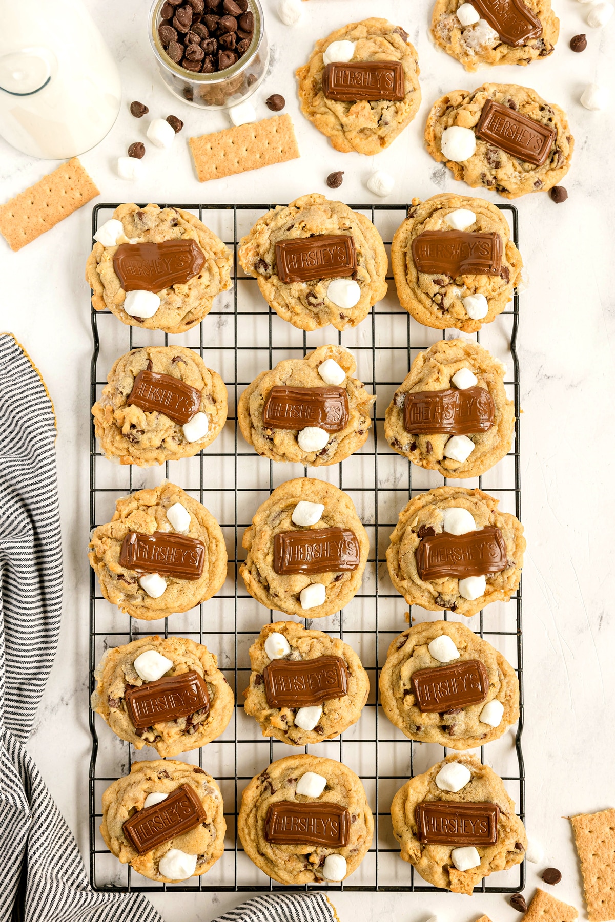 S'mores Cookies topped with melted Hershey's chocolate and mini marshmallows are cooling on a wire rack, surrounded by chocolate chips, graham crackers, and a glass of milk.