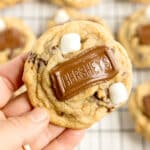 A hand holds S'mores Cookies topped with a piece of Hershey’s chocolate and mini marshmallows; more cookies are blurred in the background.