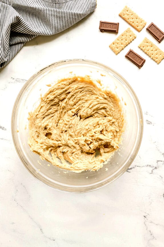 A glass bowl filled with S'mores Cookie dough sits on a white surface next to a striped cloth, chocolate pieces, and graham crackers.