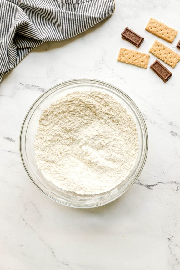 A glass bowl of flour, baking soda, and salt mixture for S'mores Cookies, sits on a white marble counter near pieces of chocolate, graham crackers, and a striped cloth napkin.