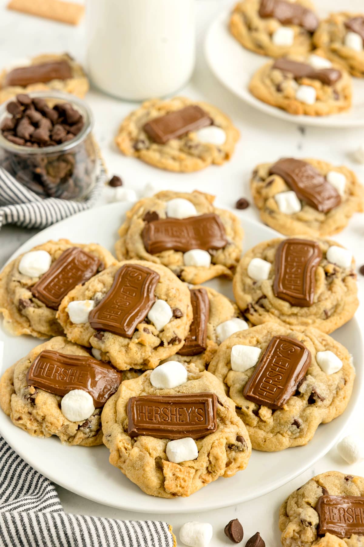 A plate of S'mores Cookies topped with chocolate pieces and mini marshmallows, with more cookies, chocolate chips, and a jar of chocolate chips in the background.