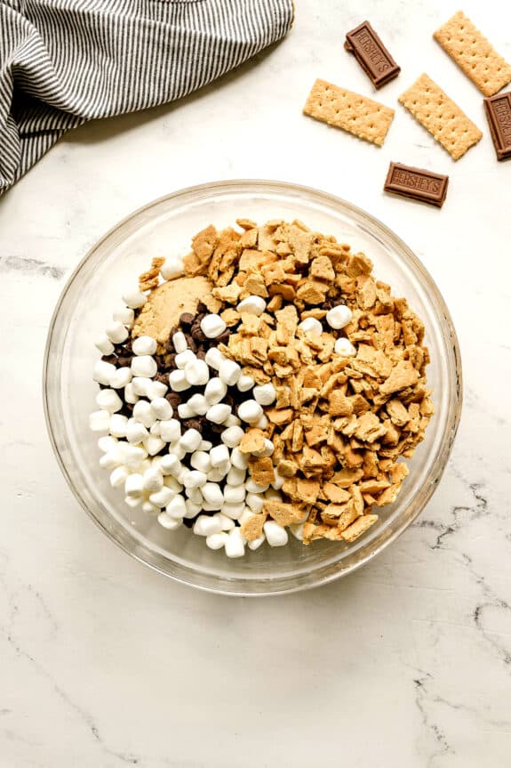 A glass bowl filled with mini marshmallows, chocolate chips, and crushed graham crackers on a white surface, with more graham crackers and chocolate pieces nearby. This is one of the steps in preparing S'mores Cookies