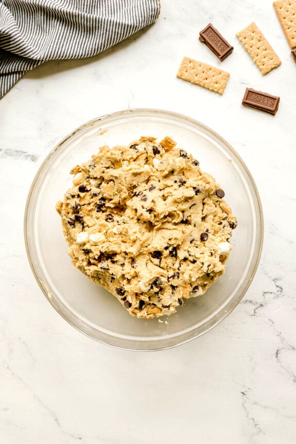 A glass bowl filled with chocolate chip cookie dough for S'mores Cookies recipe sits on a marble surface, with graham crackers, chocolate pieces, and a striped cloth nearby.