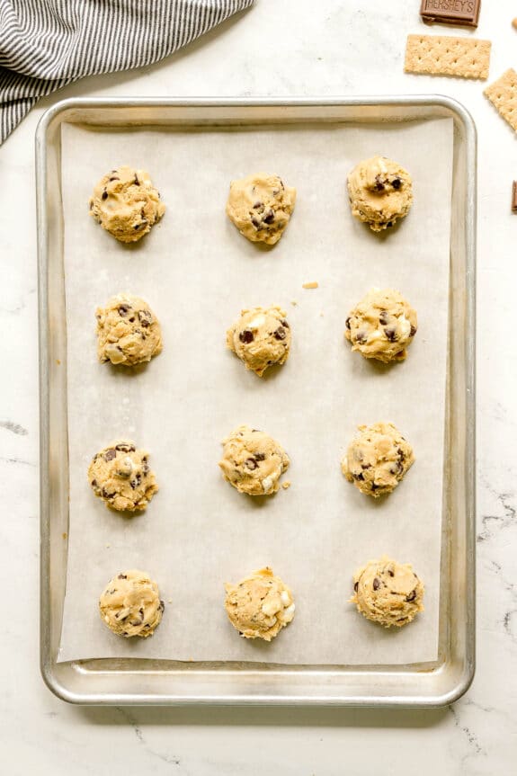 Twelve unbaked pieces of S'mores Cookies dough balls with chocolate chips on a parchment-lined baking sheet, ready for the oven.
