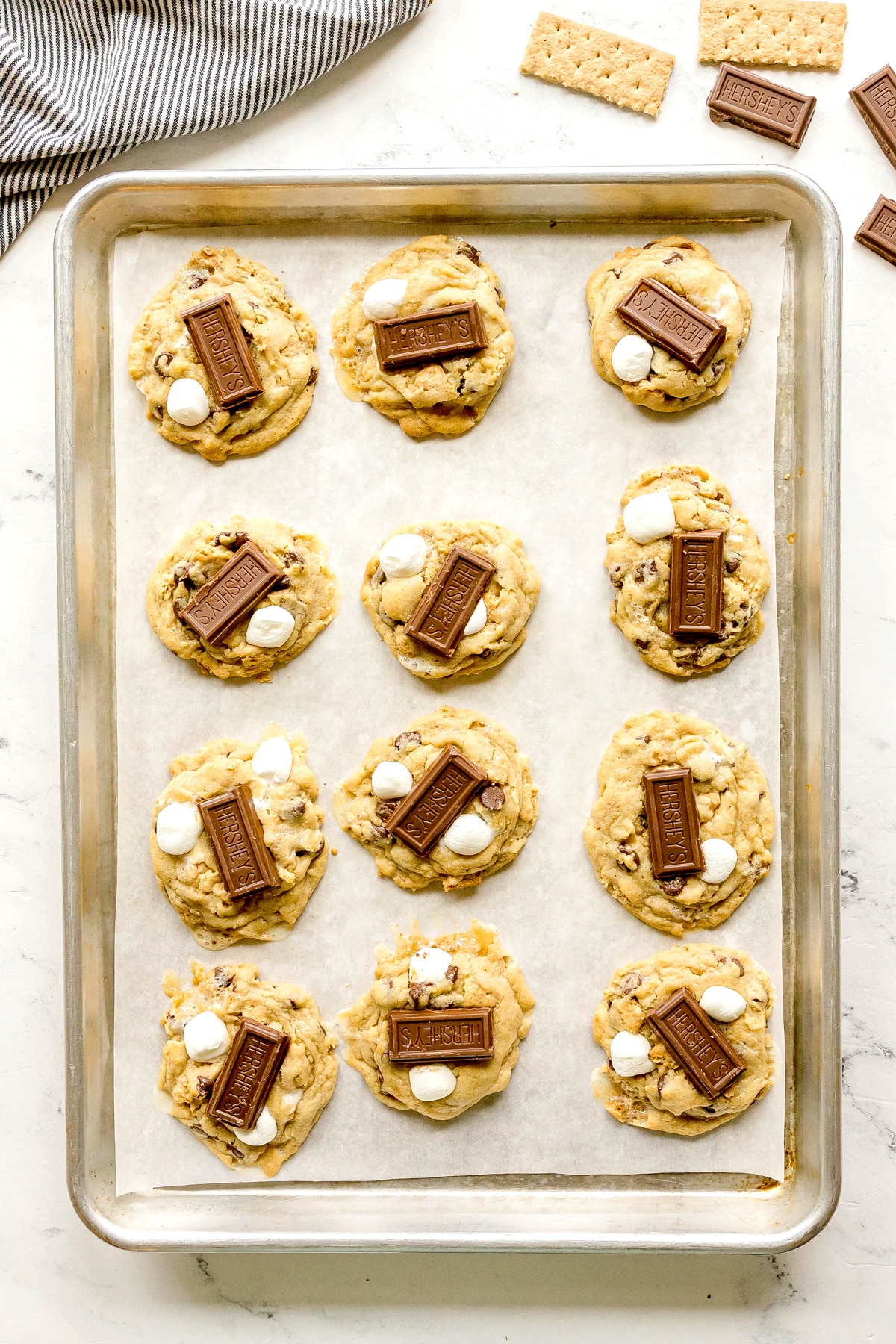 A baking sheet with twelve S'mores Cookies topped with Hershey’s chocolate pieces and mini marshmallows, on parchment paper. Some graham crackers are nearby on a white countertop.