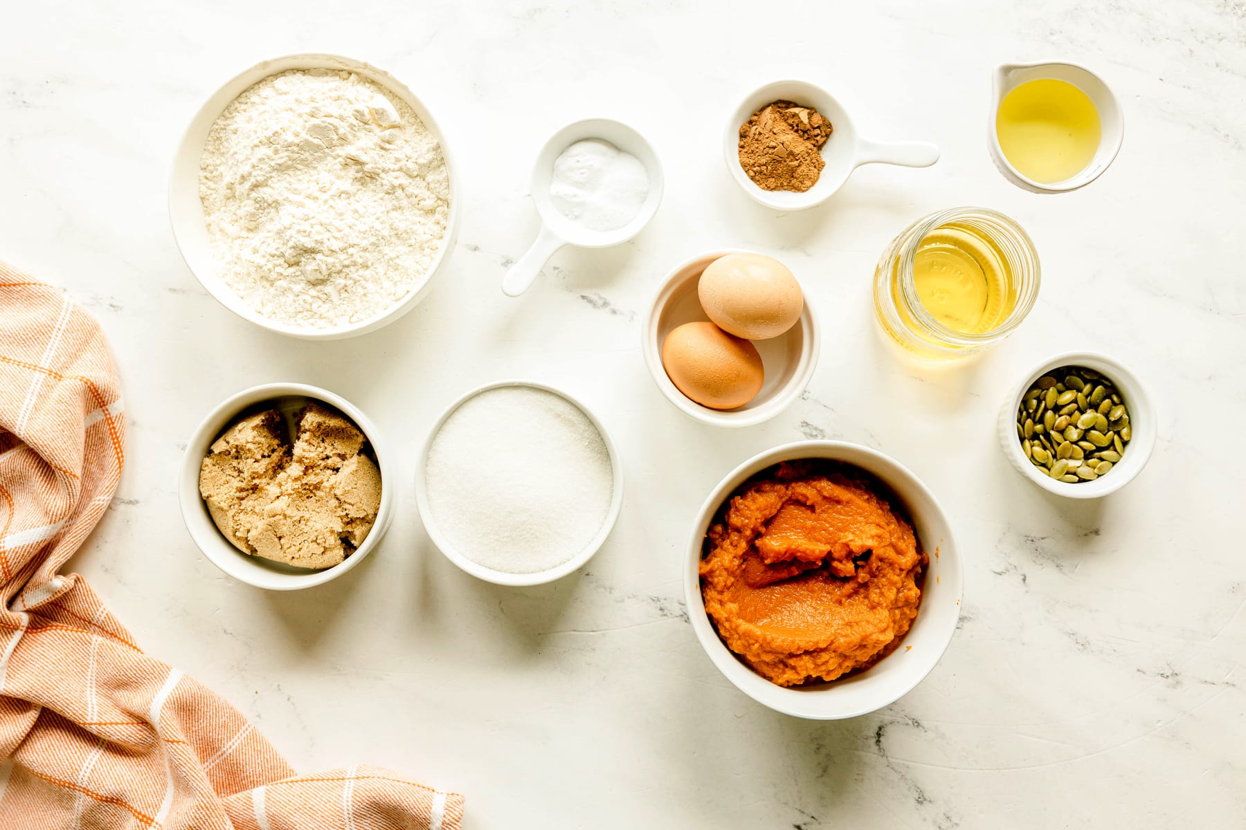 Ingredients for Starbucks Pumpkin Bread include flour, baking soda, salt, pumpkin spice mix, sugar, brown sugar, vegetable oil, eggs, apple juice, canned pumpkin, pepitas or pumpkin seeds, arranged on a white surface with a plaid towel.