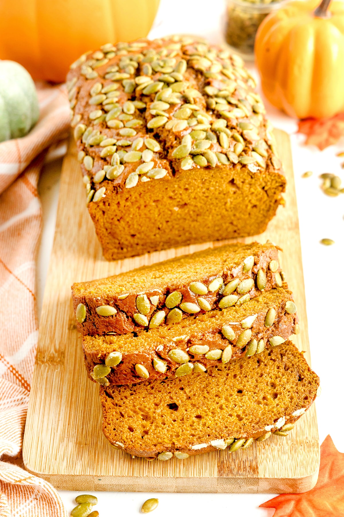 A loaf of Starbucks Pumpkin Bread topped with pumpkin seeds is partially sliced on a wooden board, surrounded by small pumpkins and a plaid cloth.
