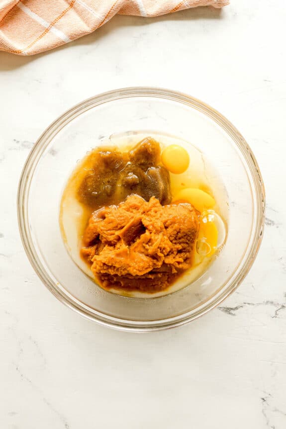 A glass bowl containing oil, eggs, apple juice, and pumpkin ingredient sits on a white marble surface next to a checkered cloth for Starbucks Pumpkin Bread