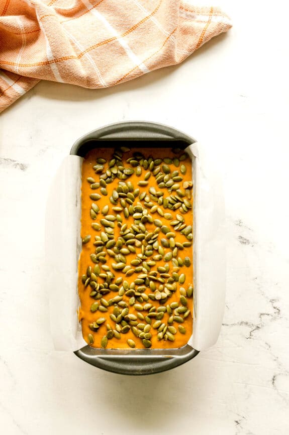 A loaf pan lined with parchment paper filled with Starbuck's Pumpkin Bread batter, topped with pumpkin seeds, sits on a white surface next to an orange checked cloth.
