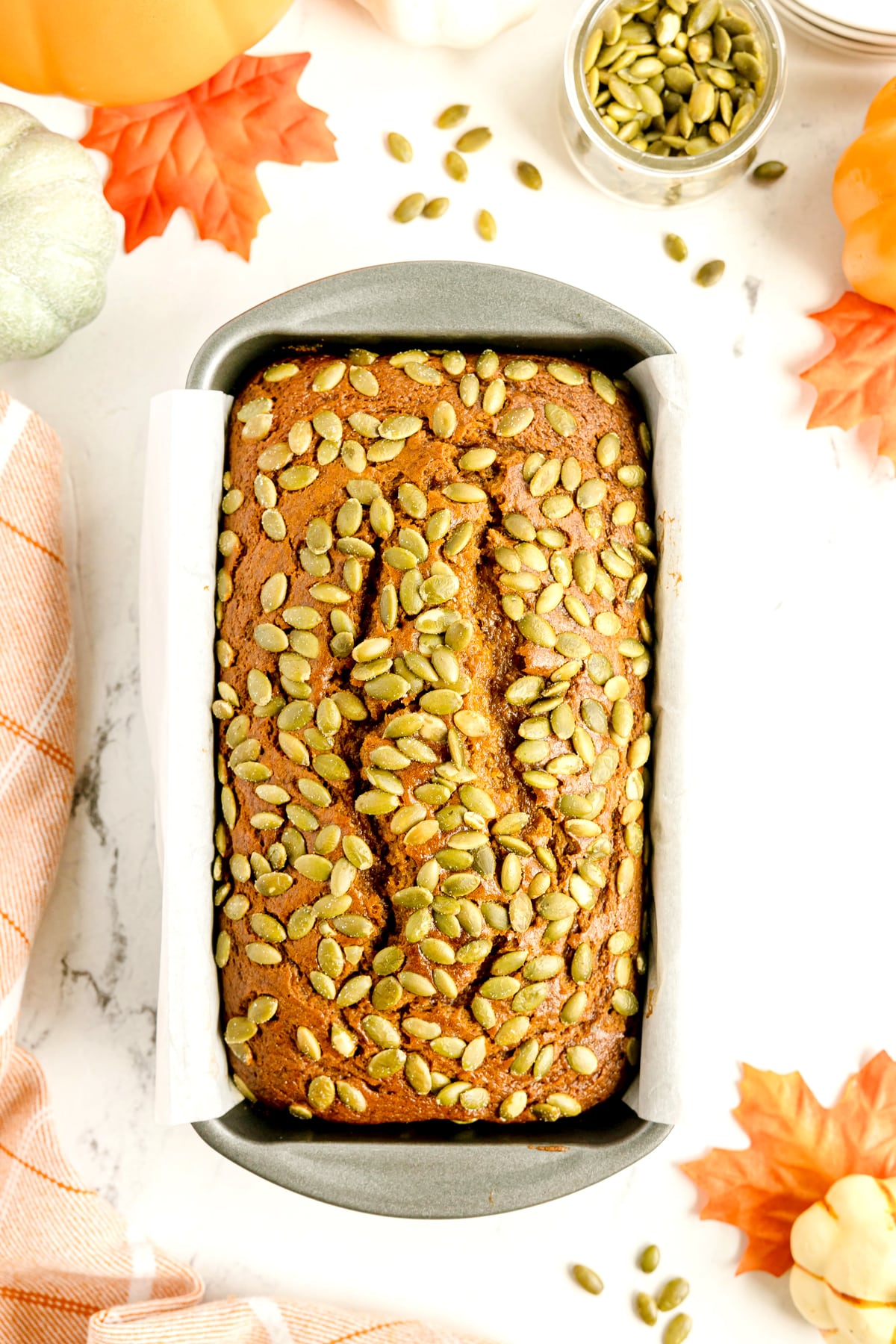 Freshly-baked Starbuck's Pumpkin Bread topped with pumpkin seeds sits in a parchment-lined baking pan, surrounded by fall leaves and pumpkin decor.
