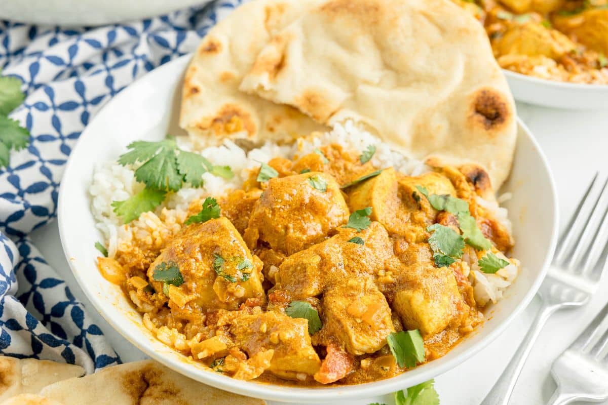 A bowl of chicken masala curry served over white rice, garnished with cilantro, and accompanied by naan bread.