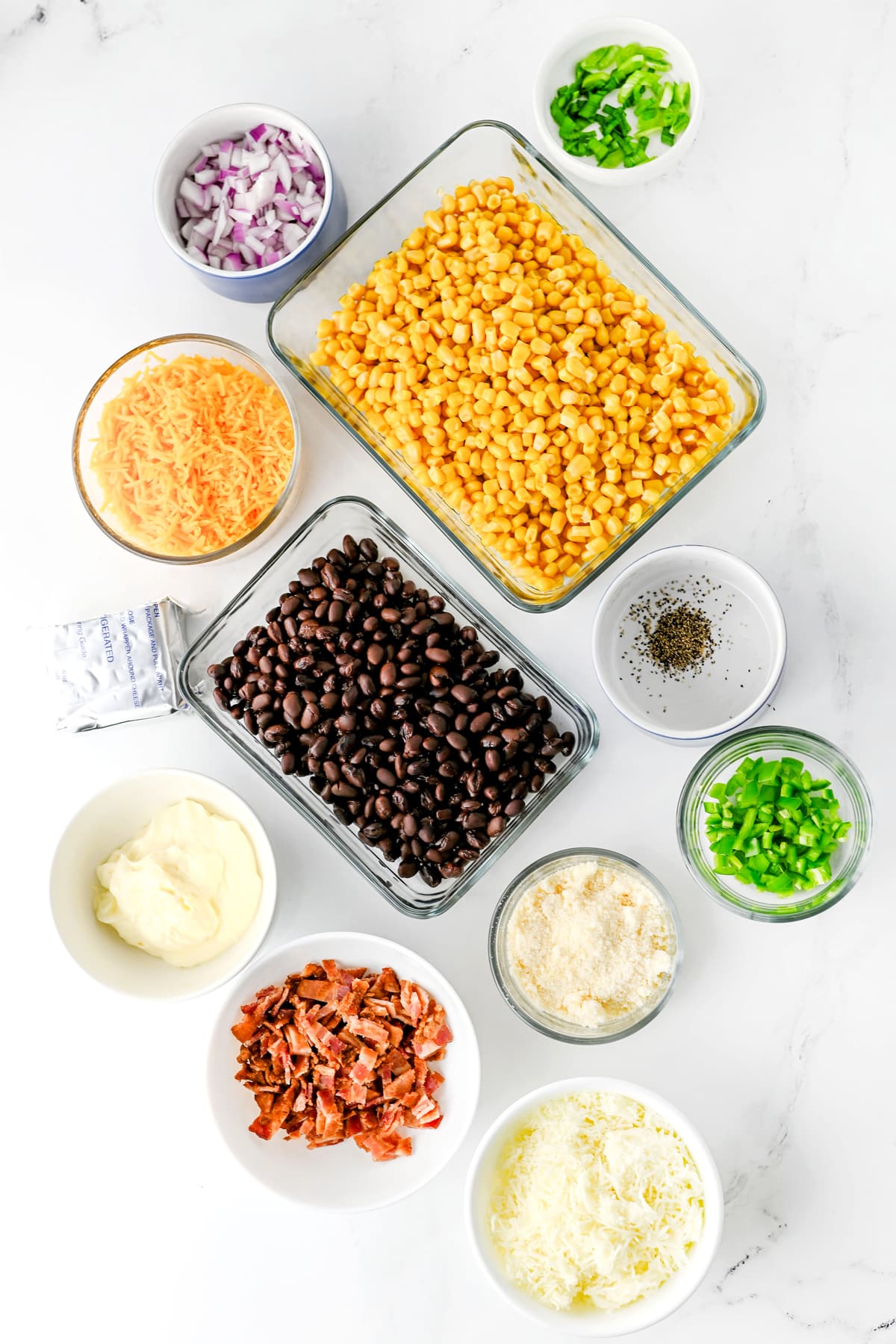 Various ingredients for Cowboy Corn Dip in bowls include whole kernel corn, black beans, cream cheese, mayonnaise, cheddar cheese, mozarella cheese, pamesan cheese, Jalapeño, bacon, red onion, black pepper and green onions.