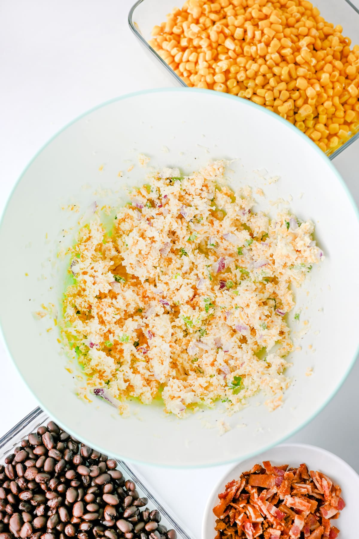 A white bowl with a mixed softened cream cheese and mayonnaise , surrounded by dishes of corn, black beans, and chopped bacon on a white surface for Cowboy Corn Dip recipe.