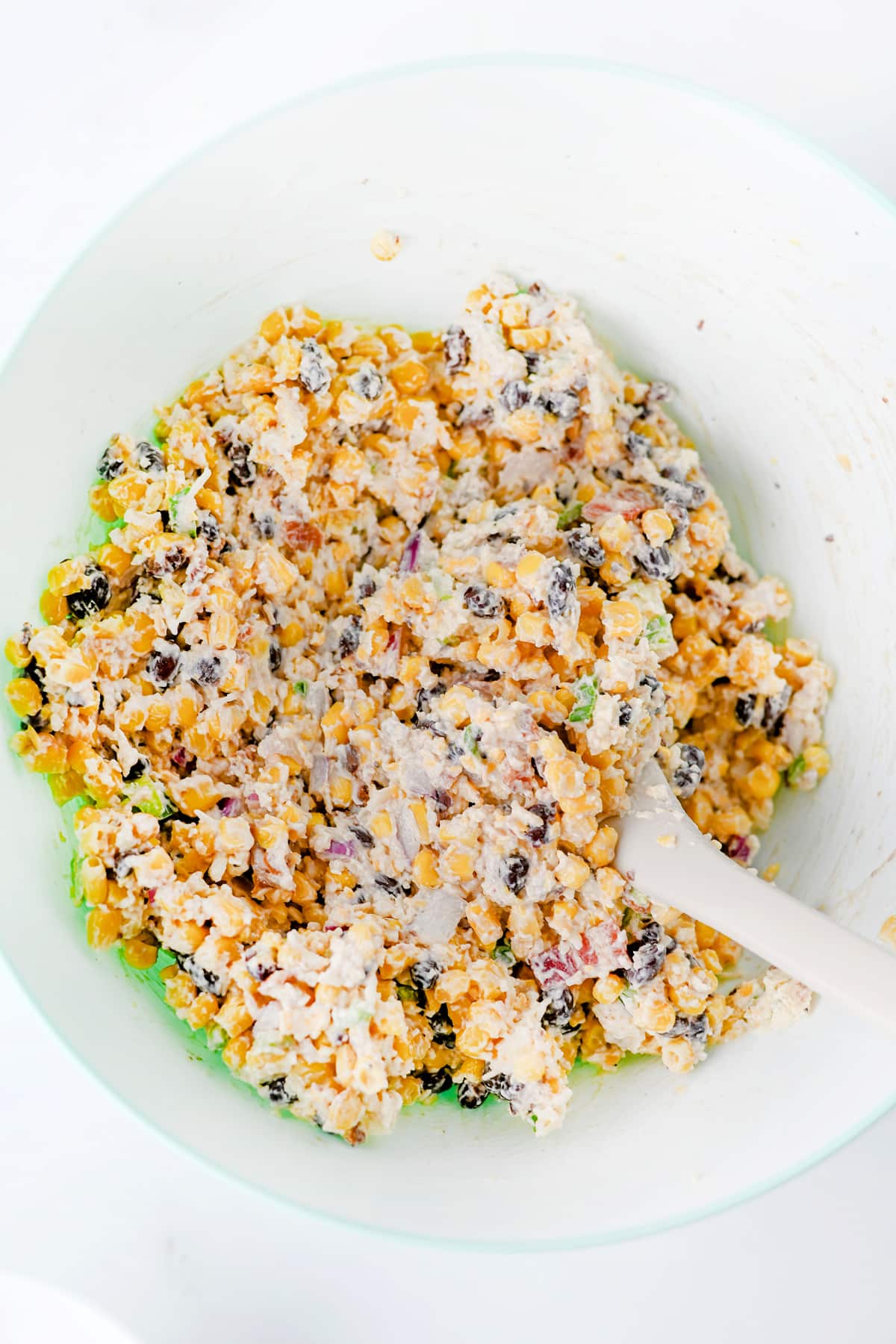 Next step in preparing Cowboy Corn Dip is to use a white mixing bowl and add into the mixture cheddar cheese, mozzarella cheese, parmesan cheese, jalapeño, red onion, and black pepper. There is also a white spatula inside.