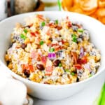 A white bowl filled with Cowboy Corn Dip or a mixed salad containing bacon, black beans, corn, red onions, green onions, and a creamy dressing.