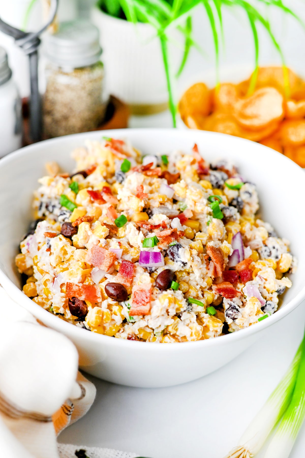 A white bowl filled with Cowboy Corn Dip containing corn, beans, bacon, chopped red onion, and green onions, set on a white surface with ingredients in the background.