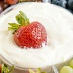 A whole strawberry partially dipped in creamy white fruit dip recipe, with grapes and blueberries blurred in the background.