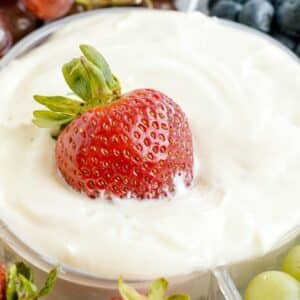 A whole strawberry partially dipped in creamy white fruit dip recipe, with grapes and blueberries blurred in the background.