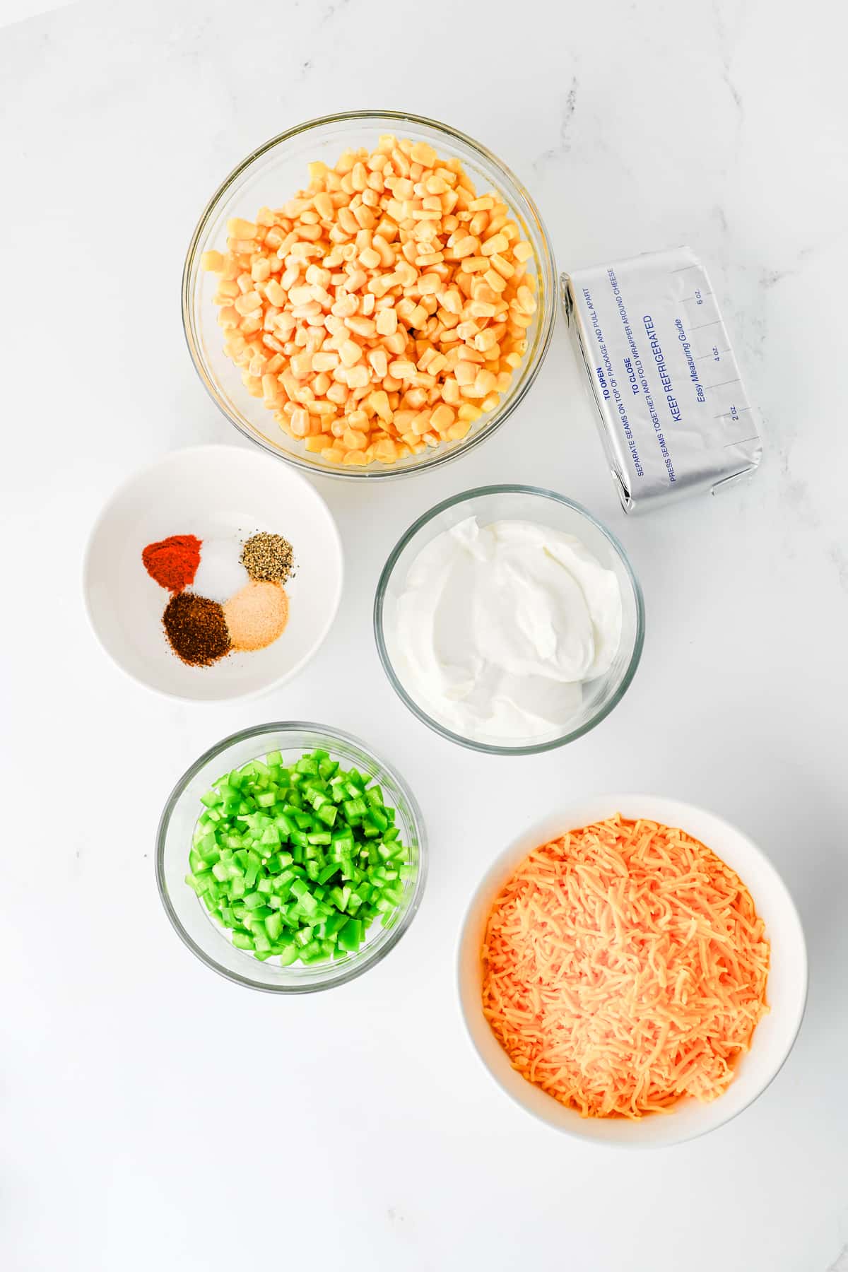 Top view of bowls containing ingredients for Jalapeno Corn Dip are cream cheese, sour cream, jalapenos, cheddar cheese, chilli powder, garlic powder, salt, black pepper, and smoked paprika on a white counter.