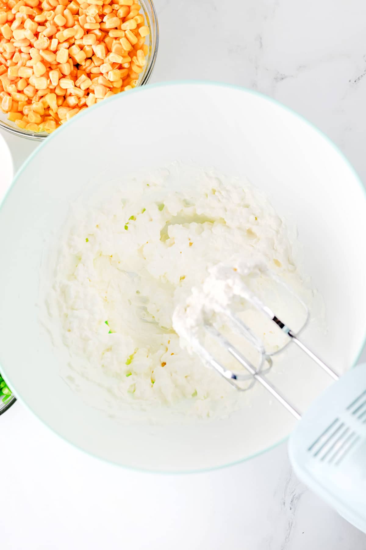 Electric hand mixer with whipping cream cheese and sour cream in a white mixing bowl, with a bowl of corn kernels visible in the background on a marble countertop. This is the first step in making Jalapeno Corn Dip