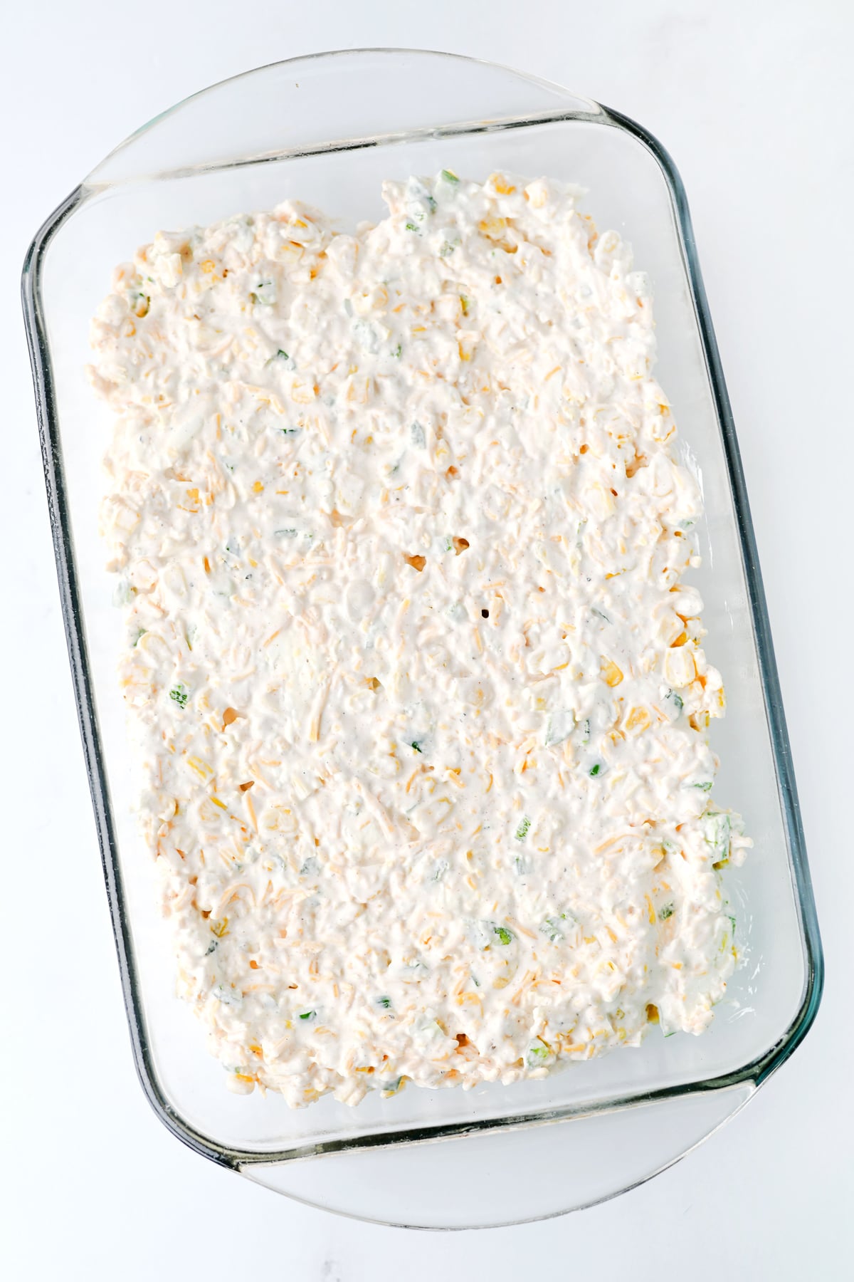 Rectangular glass baking dish filled with an uncooked, creamy, white mixture with bits of vegetables and cheese, ready for baking, on a white surface.