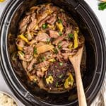 Shredded beef with onions and peppers in a slow cooker, stirred with a wooden spoon—perfect for making savory Slow Cooker Mississippi pot roast.