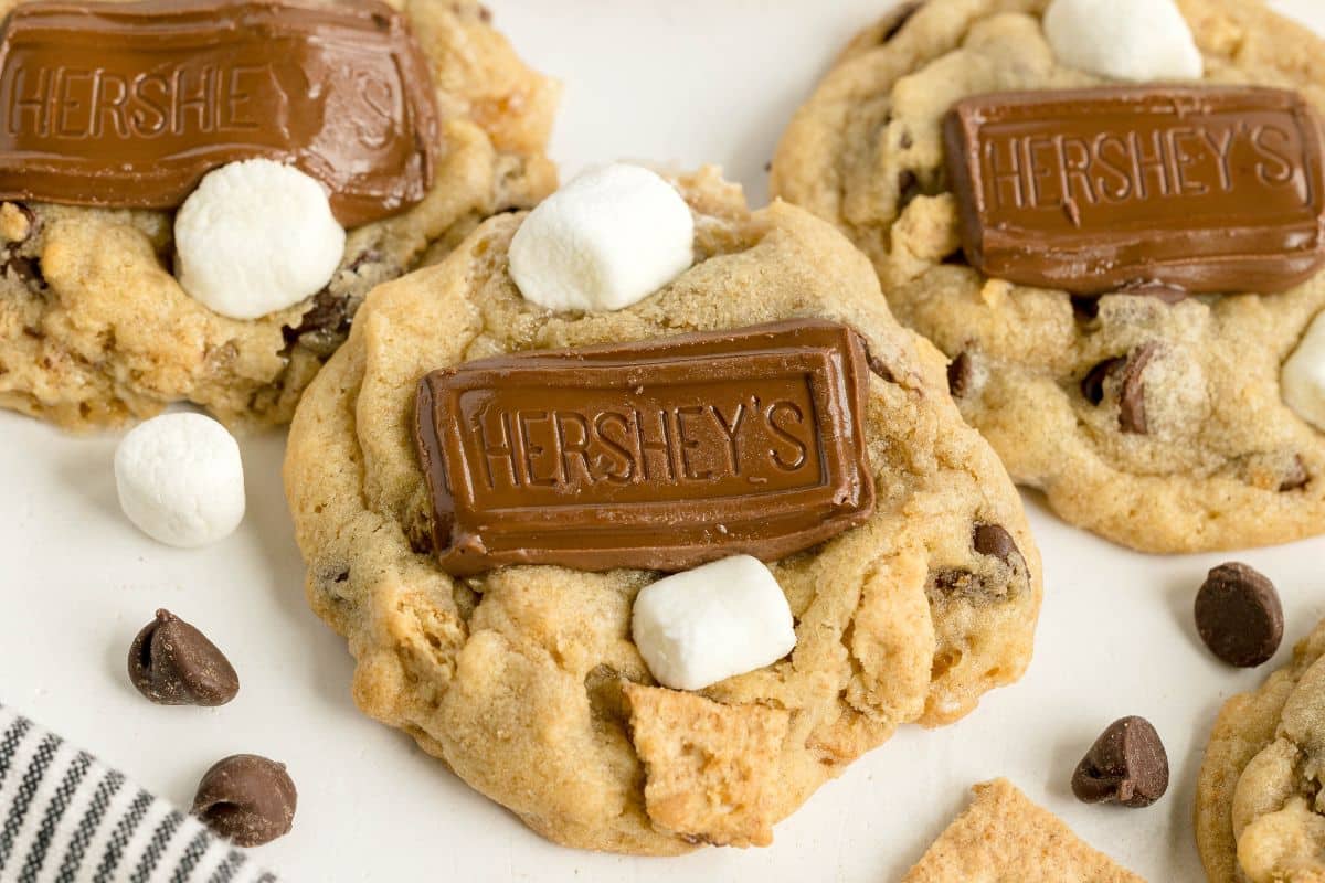 Smores Cookies: Chocolate chip cookies topped with a Hershey's chocolate piece and mini marshmallows, capturing all the classic flavors of smores, with extra chocolate chips and marshmallows scattered around on a white surface.
