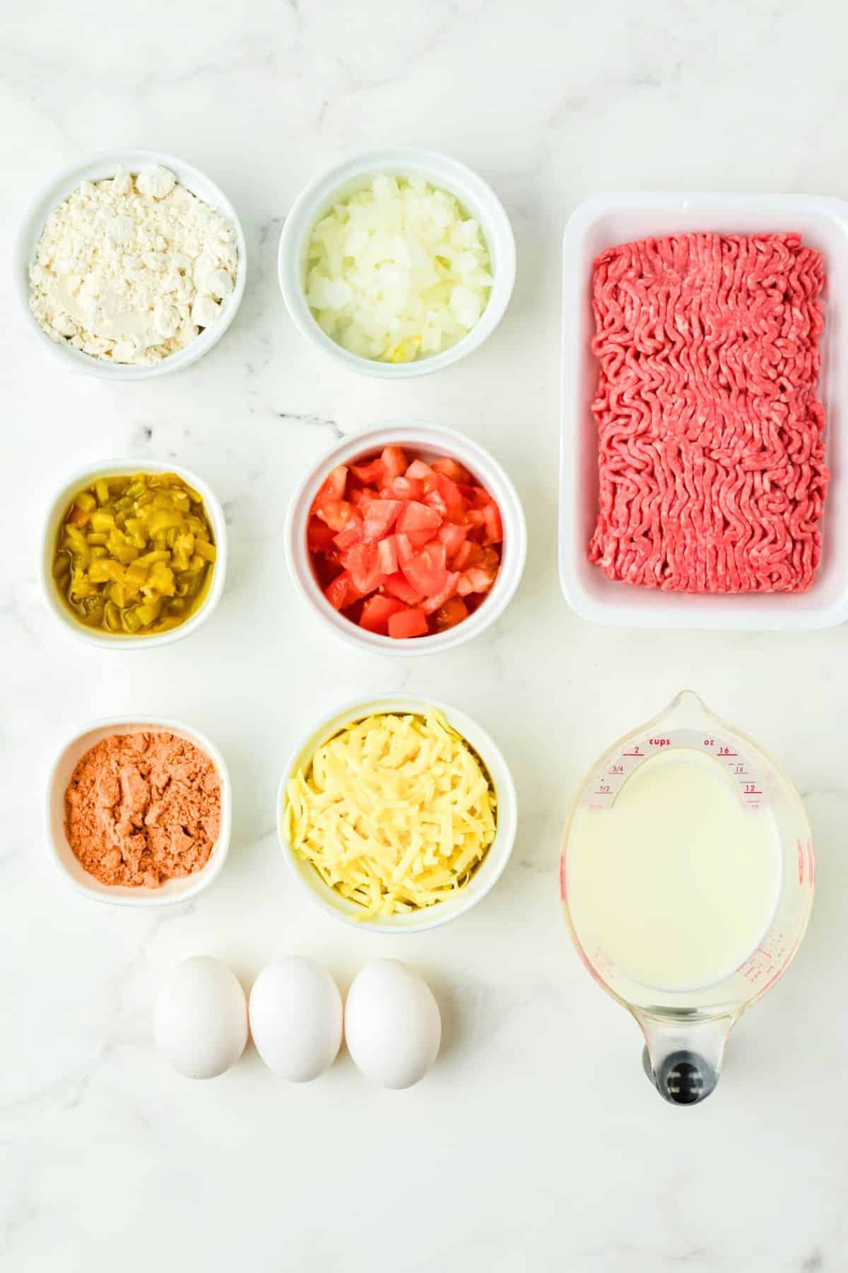 Nine ingredients for a taco pie recipe are arranged on a white surface, including ground beef, milk, eggs, shredded cheese, diced tomatoes, chopped onions, relish, a powder mix, and flour.